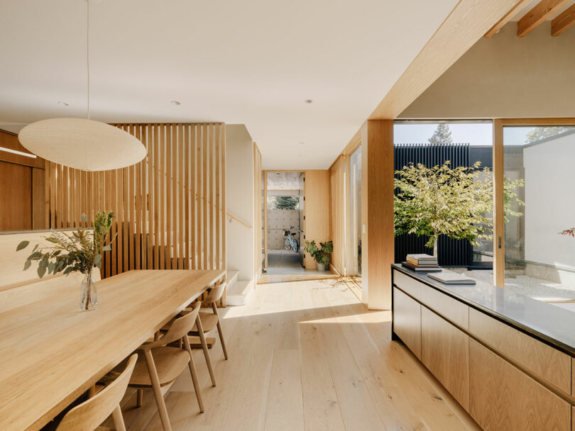 A modern dining and kitchen area with light wood furniture, large windows, and natural light, featuring a dining table, wooden slats, and a view of a small courtyard with plants.