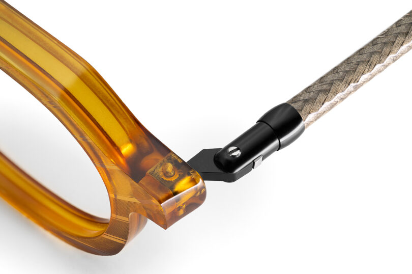 Close-up view of the hinge on a pair of eyeglasses, featuring a translucent amber frame and a textured, beige temple with a black metal connector.