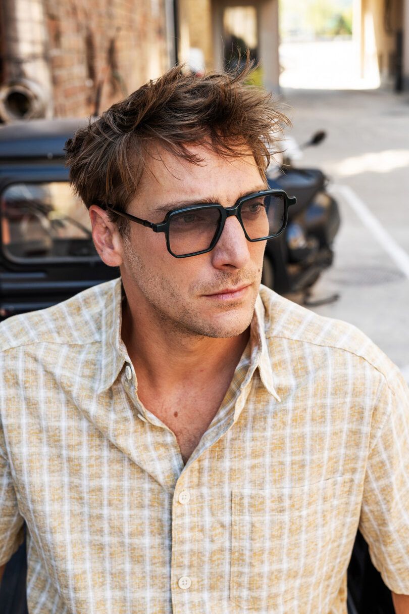 A man wearing large black glasses and a light plaid shirt stands outdoors in front of a black car and a brick wall.