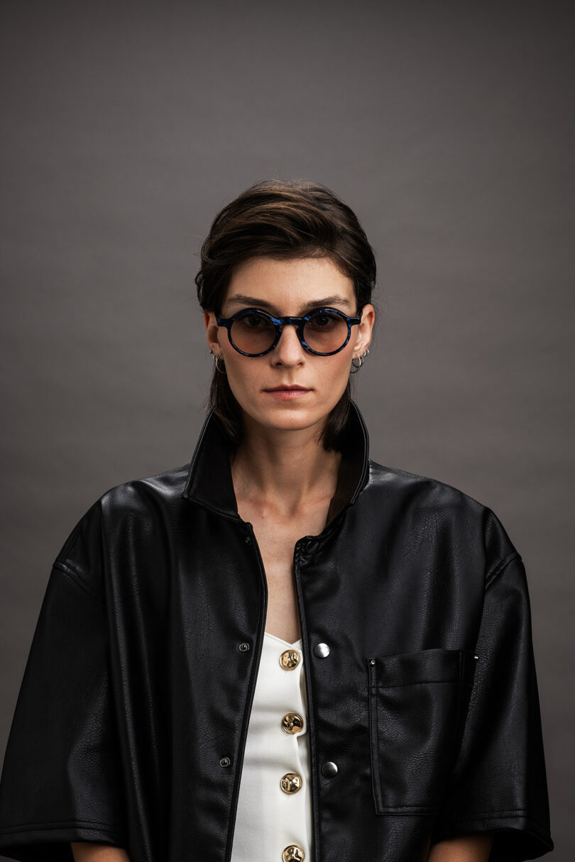 A woman with short dark hair wears round sunglasses, a black faux leather jacket with an open collar, and a white top with gold buttons, standing against a gray background.