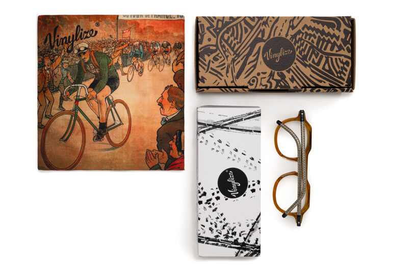 Eyeglasses with tortoiseshell frames, a patterned case, a white cleaning cloth, and a vintage-style cycling illustration branded "Vinylize".