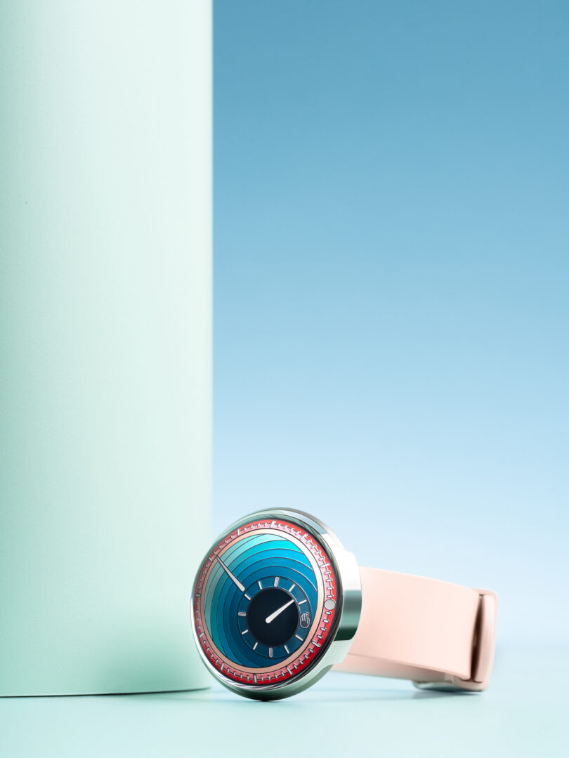 A wristwatch with a blue face, pink bezel, and light pink strap is propped against a light mint green cylindrical object on a blue background