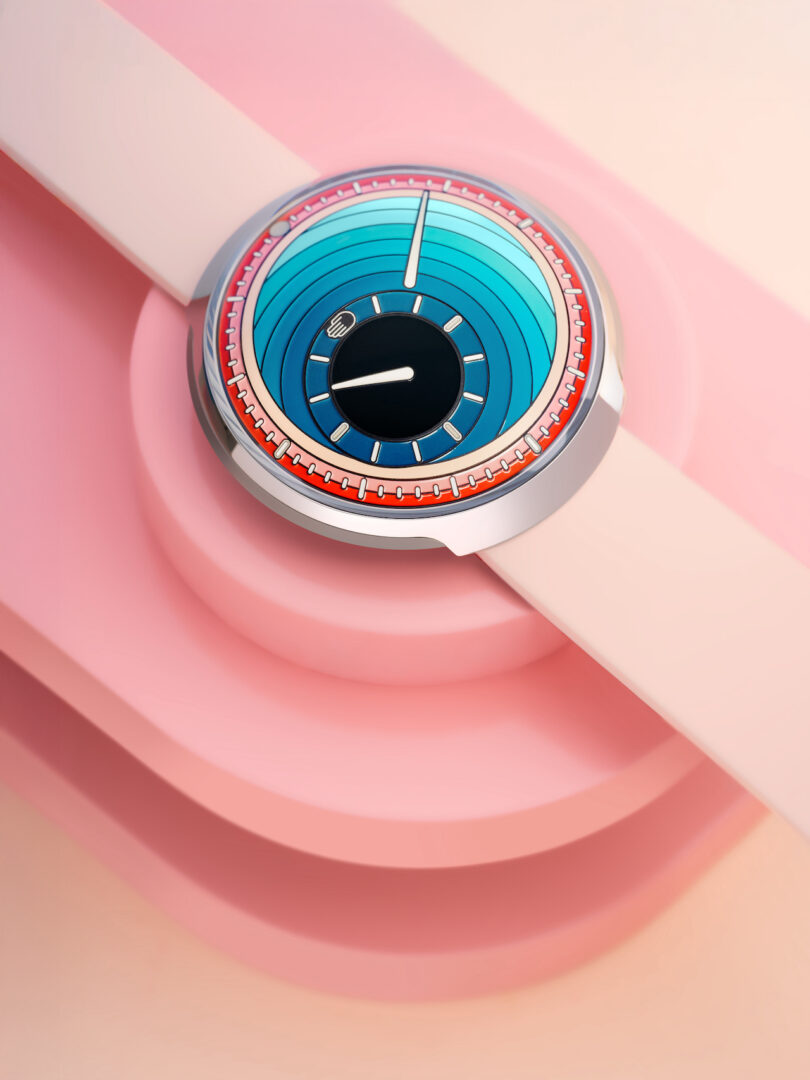 A modern wristwatch with a blue and red circular dial and a light pink strap, placed on a geometric pink background