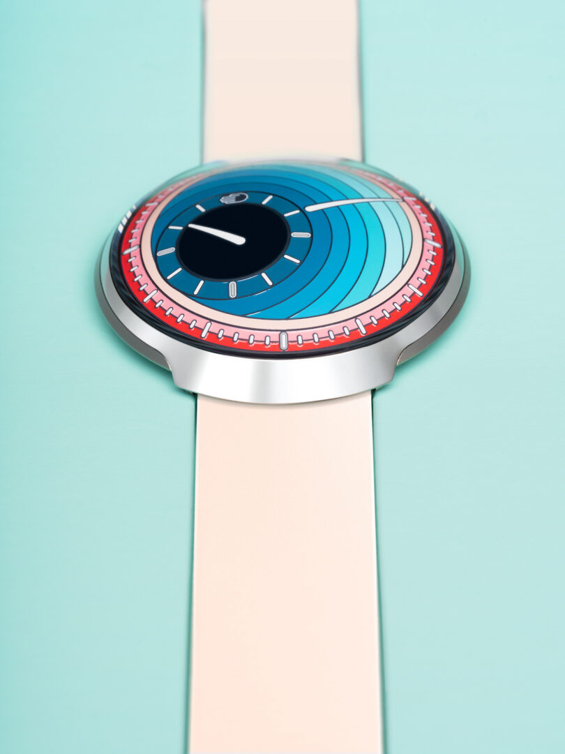 A modern wristwatch with a circular, blue and turquoise face, red detailing, and a light beige strap, displayed on a light blue background