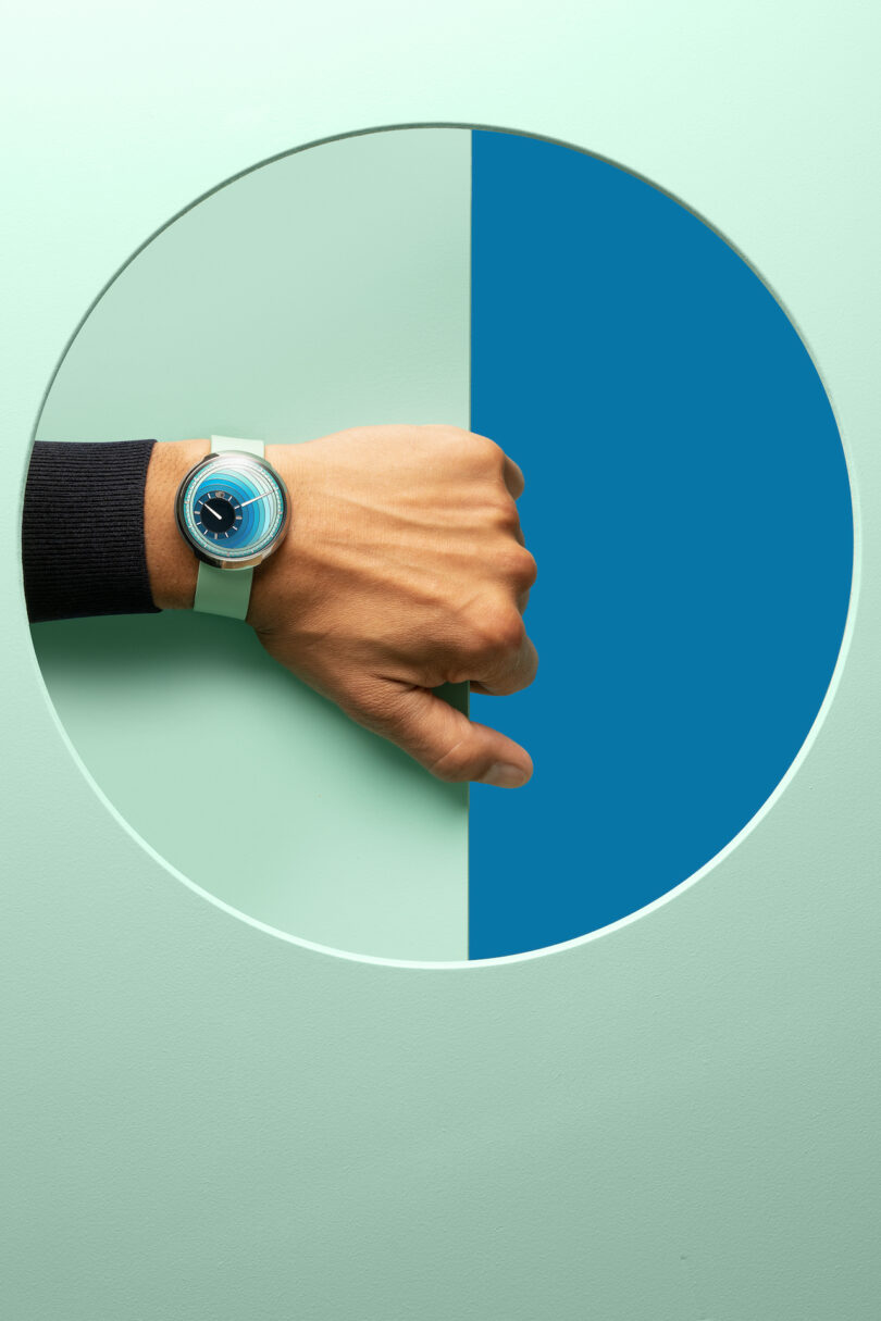 A hand wearing a light blue wristwatch is placed through a circular cutout in a wall divided into mint green and teal blue halves