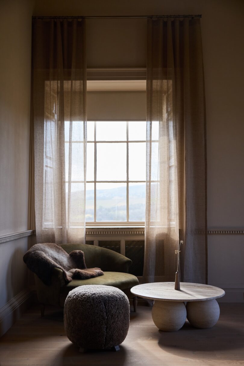 A sunlit room with sheer curtains, an olive green armchair with a fur throw, a round textured ottoman, and a round white coffee table with a vase.
