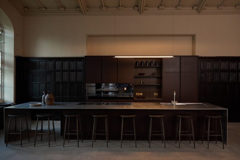 A modern, minimalist kitchen with dark cabinetry, an island with seven stools, vases, shelving, built-in appliances, and soft lighting in a high-ceilinged room.