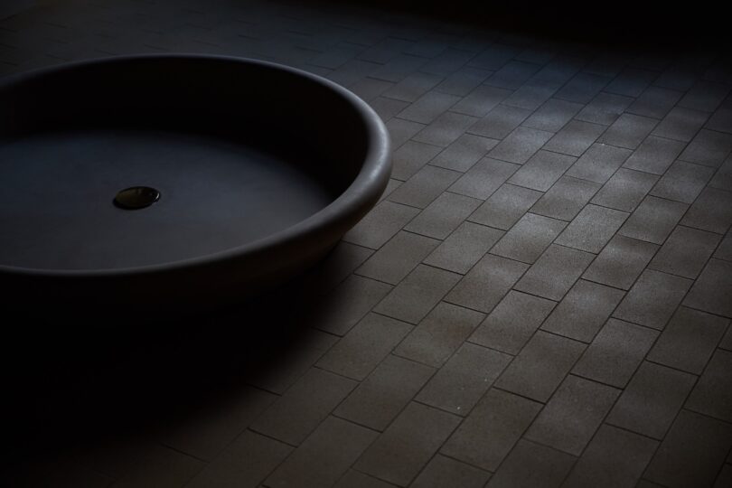 A large, round, dark basin sits on a floor made of rectangular brown tiles, with light falling unevenly across the scene.