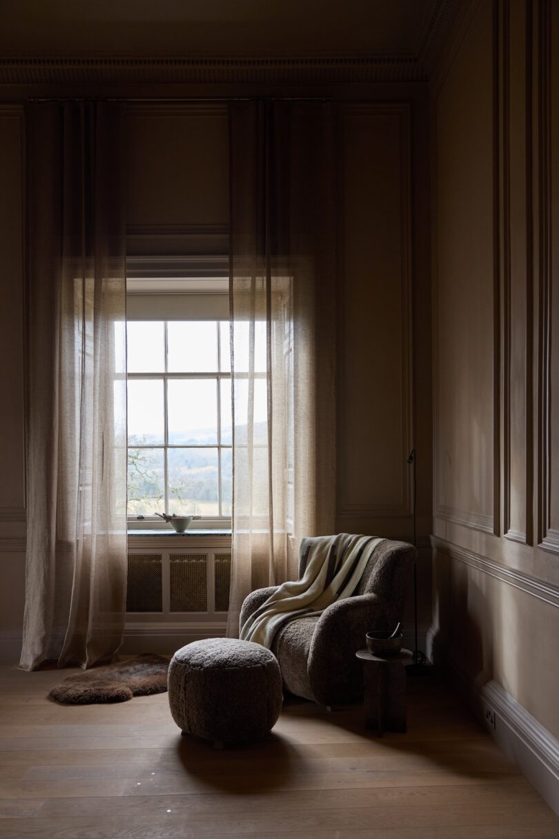 A cozy armchair with a blanket, a round ottoman, and a small side table sit by a large window with sheer curtains, letting in natural light in a minimalist room.
