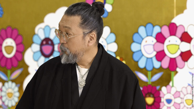 A man with a beard, glasses, and a black robe stands in front of a gold background decorated with colorful, smiling cartoon flowers.