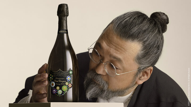 A man with glasses and gray hair tied in a bun examines a decorated bottle of Dom Pérignon champagne placed on a white surface.