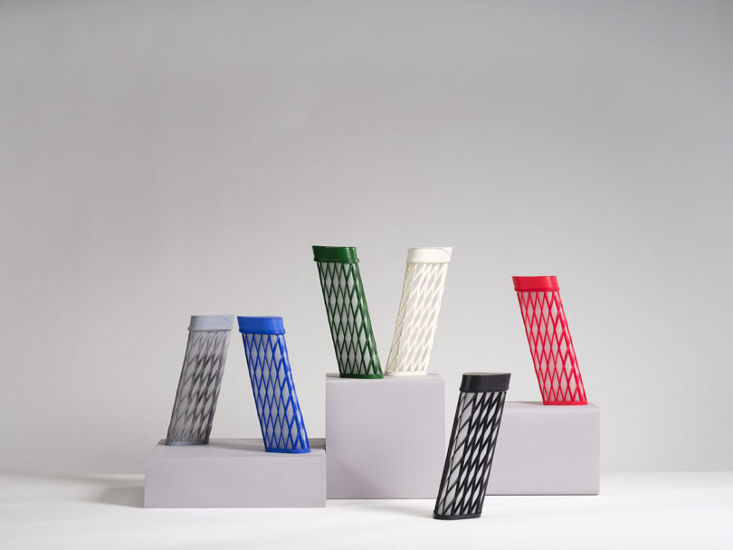Six cylindrical containers with diamond-patterned mesh exteriors in various colors are displayed on white rectangular blocks against a plain background.