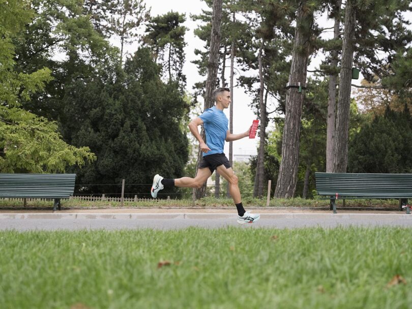 A man in athletic wear runs with a water bottle in a park, passing two empty green benches and tall trees in the background.