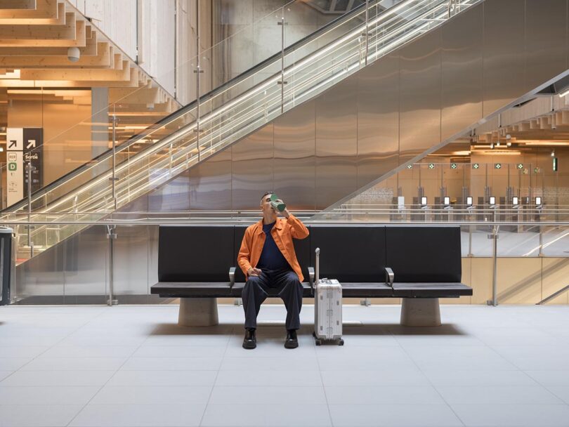 A man in an orange jacket sits on a bench with a suitcase, drinking from a bottle in a modern building with metal stairs in the background.
