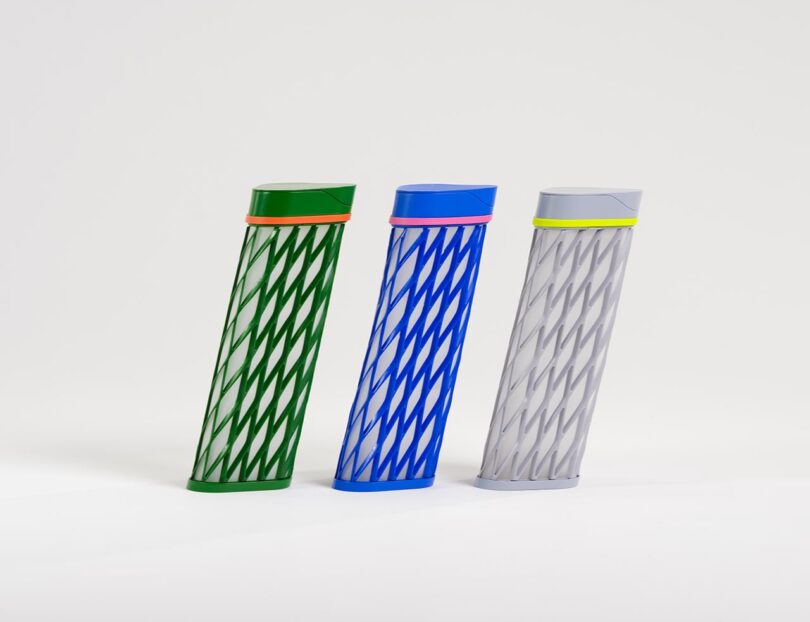 Three cylindrical objects with diagonal cut-out patterns in green, blue, and gray, each with a contrasting colored band near the top, standing upright on a plain white background.