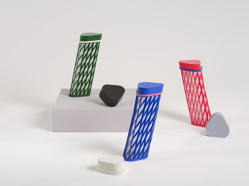 Five colorful, geometric containers with crisscross patterns stand on and around a rectangular platform against a plain white background.