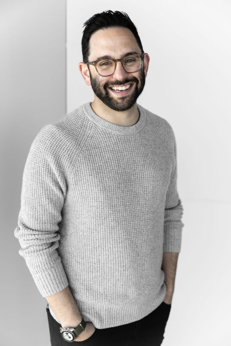 A man with dark hair and glasses, wearing a gray knit sweater and black pants, stands against a light background with his hands in his pockets, smiling at the camera.