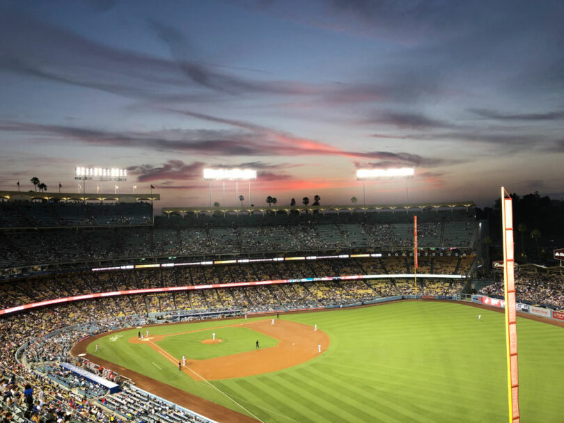 A baseball game underway at a stadium in the evening with the sky showing sunset colors and stadium lights illuminating the field.