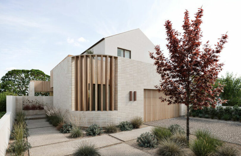 Modern two-story house with light brick walls, wood accents, and minimalist landscaping featuring a red-leafed tree and various grasses in the front yard.