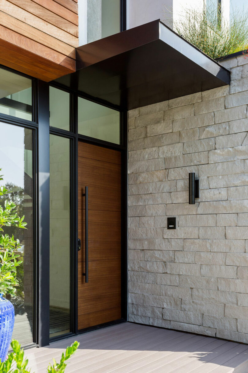 Modern house entryway with a wooden door, black handle, stone wall, glass panels, and a flat black metal awning overhead.