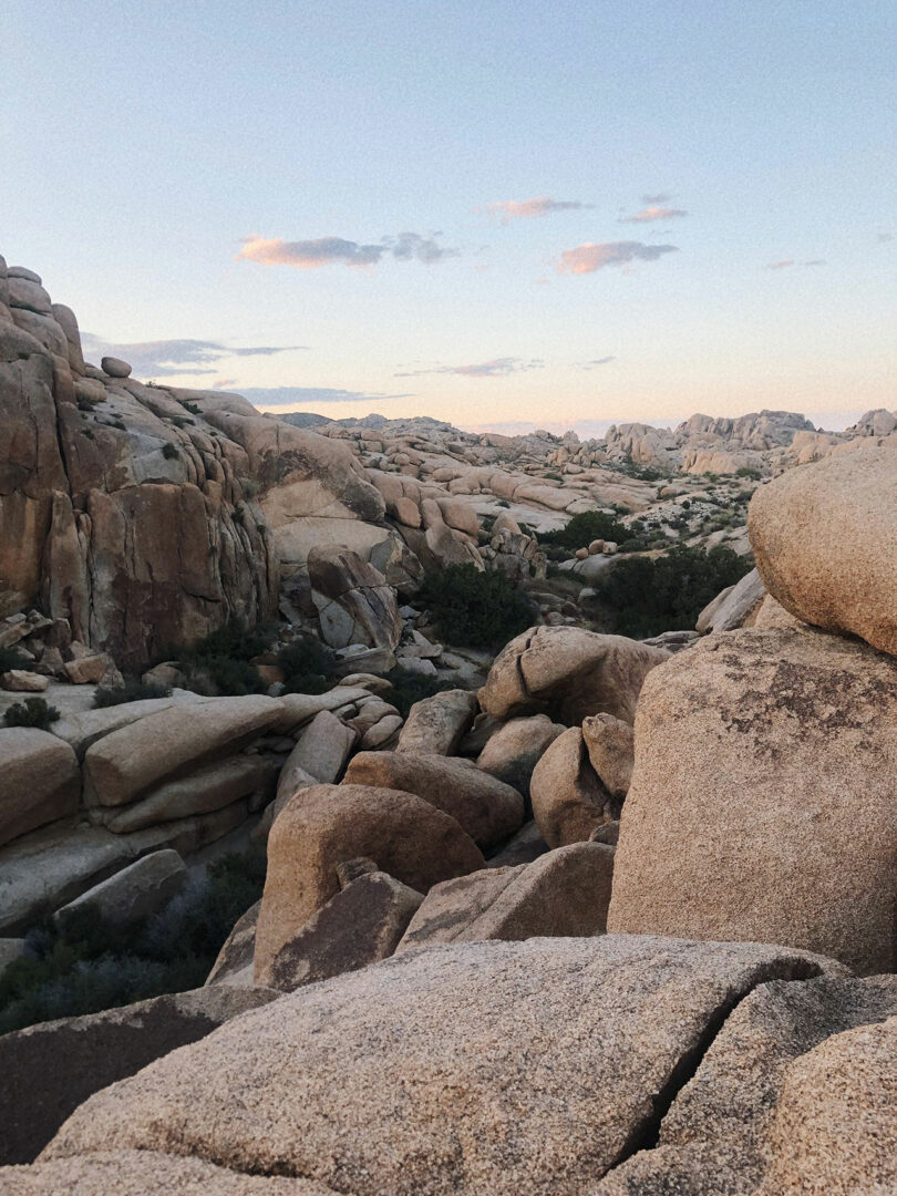 Large rock formations and scattered boulders under a clear sky with a few clouds, surrounded by sparse vegetation in a desert landscape during sunset.
