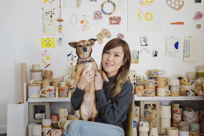 A woman sits in a pottery studio, holding a small brown dog. Ceramic mugs and abstract art are displayed on shelves and walls behind her.