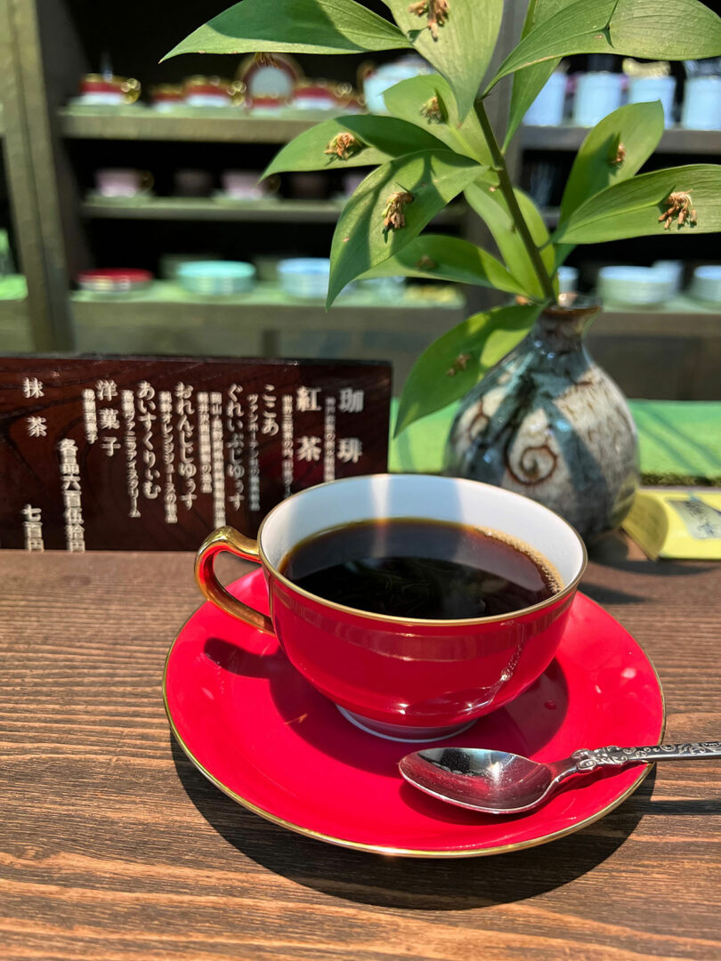 A cup of black coffee in a red cup and saucer sits on a table with a spoon, a vase with green leaves, and a Japanese menu in the background.