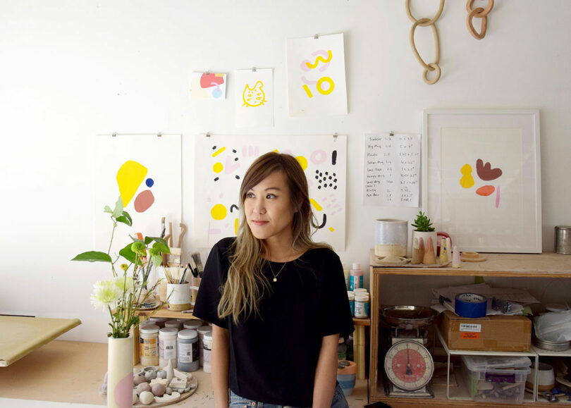 A woman stands in an art studio with colorful abstract artworks on the wall, surrounded by art supplies, plants, and ceramics.