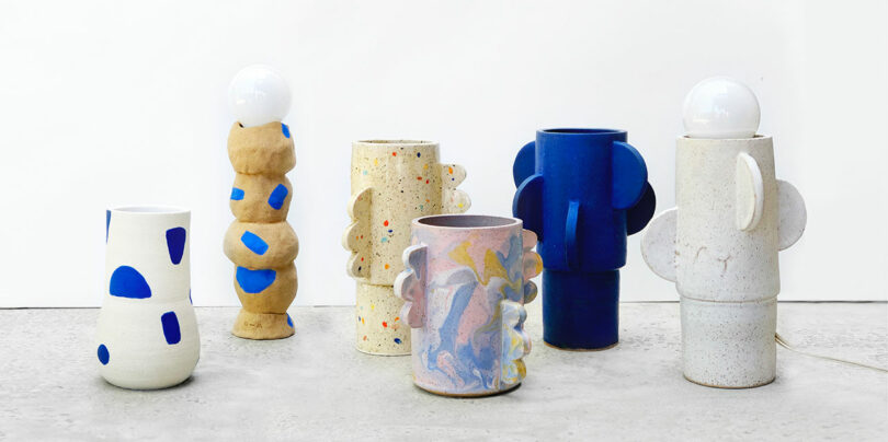 Six uniquely shaped ceramic vases and lamps with abstract patterns and blue, beige, and multicolor designs are displayed on a concrete surface against a white background.