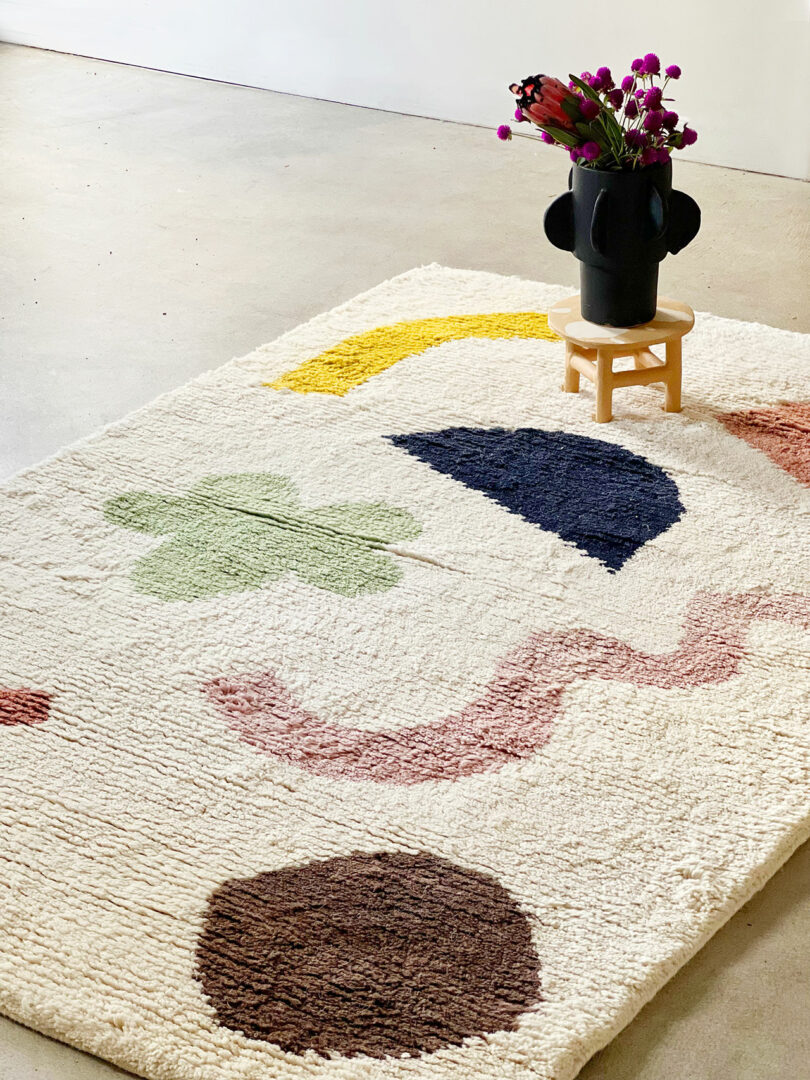 A white rug with colorful abstract shapes lies on a concrete floor. A small wooden stool with a black vase holding pink flowers sits on the rug.