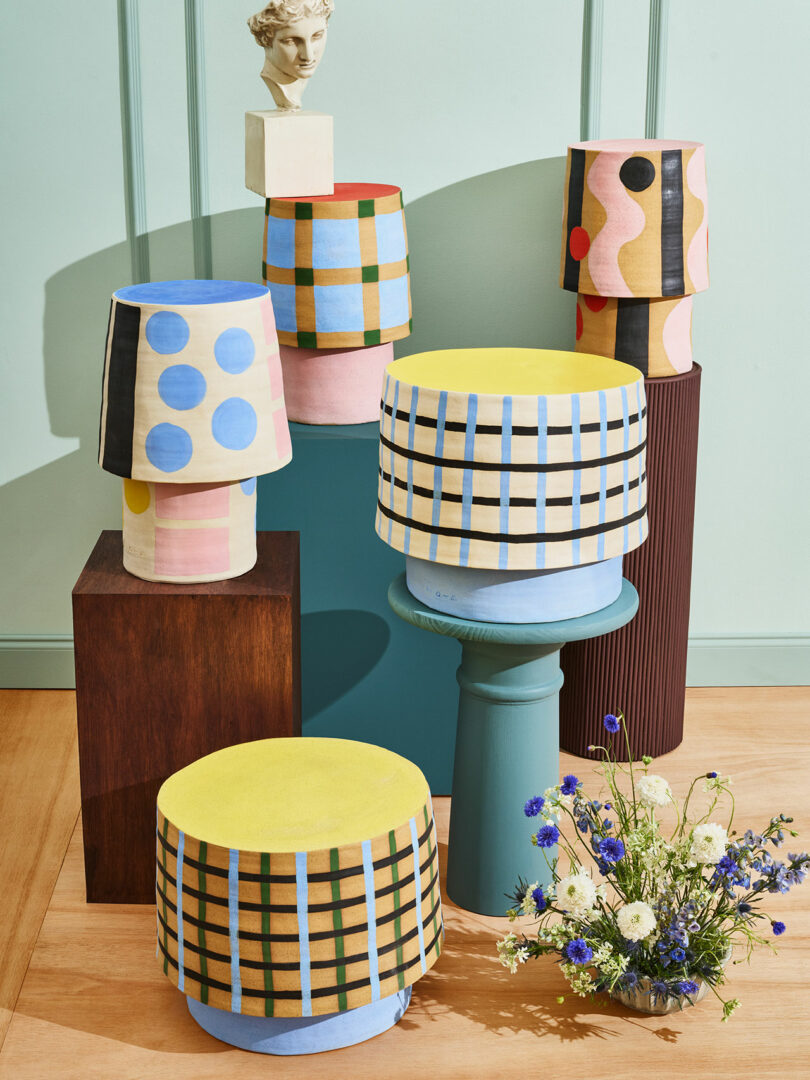 A group of stools with colorful geometric patterns are arranged on platforms; a small bust and a vase of wildflowers are also displayed.