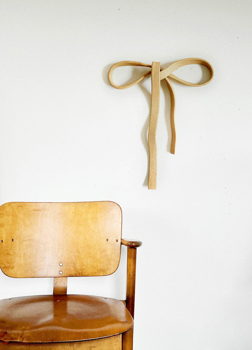 A wooden chair is positioned against a white wall with a tan leather strap tied in a bow and hung above it.