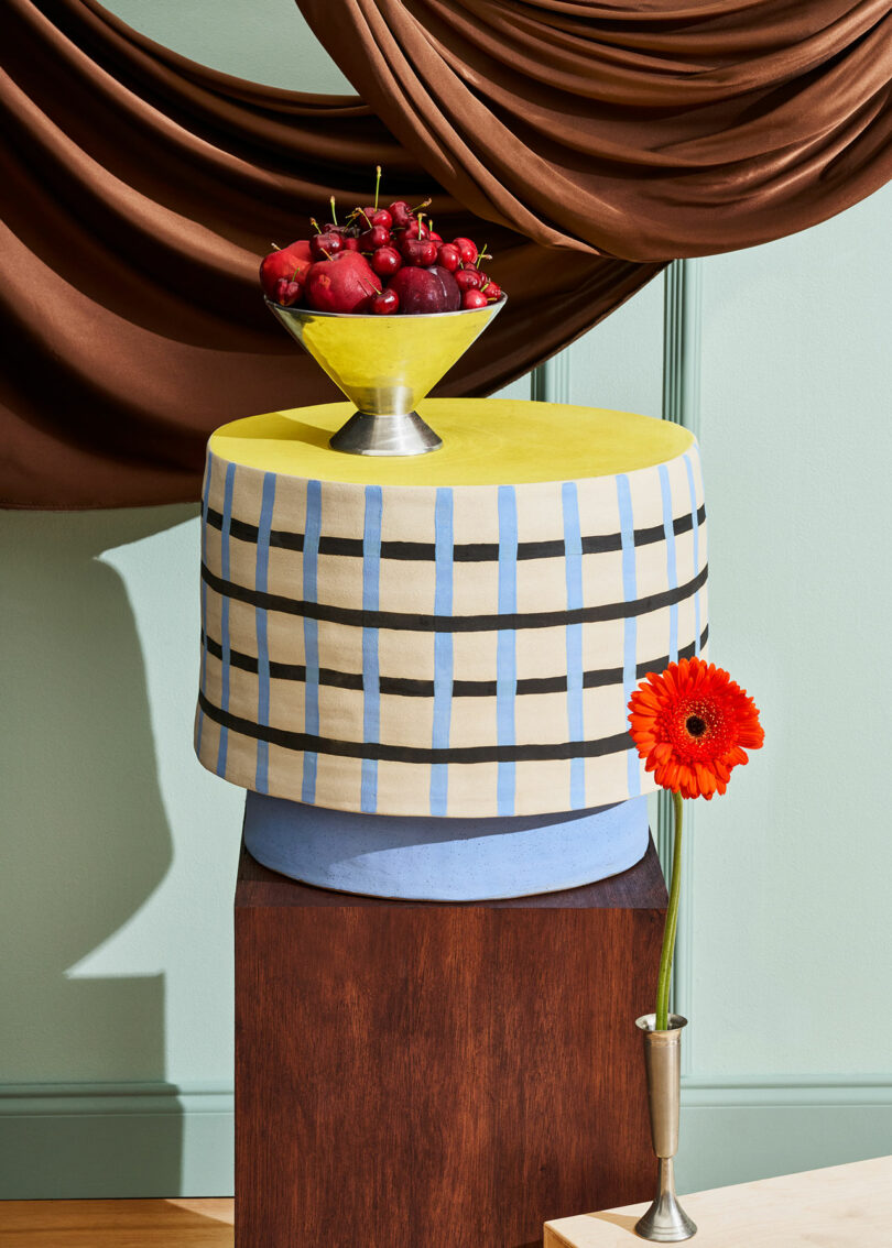 A yellow and blue checkered round table with a fruit bowl of red apples and cherries on top, next to an orange gerbera daisy in a small vase, with brown draped fabric in the background.