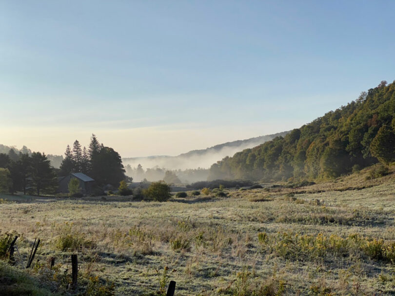 A frosty meadow with morning mist, bordered by wooded hills, under a clear sky; a barn stands near trees on the left.