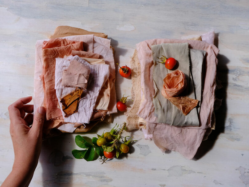 A hand reaches toward neatly stacked pieces of pastel-colored fabric, with small tomatoes and green leaves arranged between the stacks on a light wooden surface.