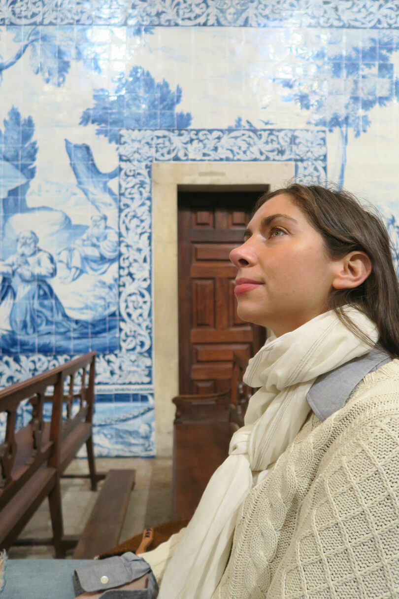 Kiva Motnyk sits on a bench inside a building, wearing a white scarf. Blue and white tile art and a wooden door provide an artistic backdrop.