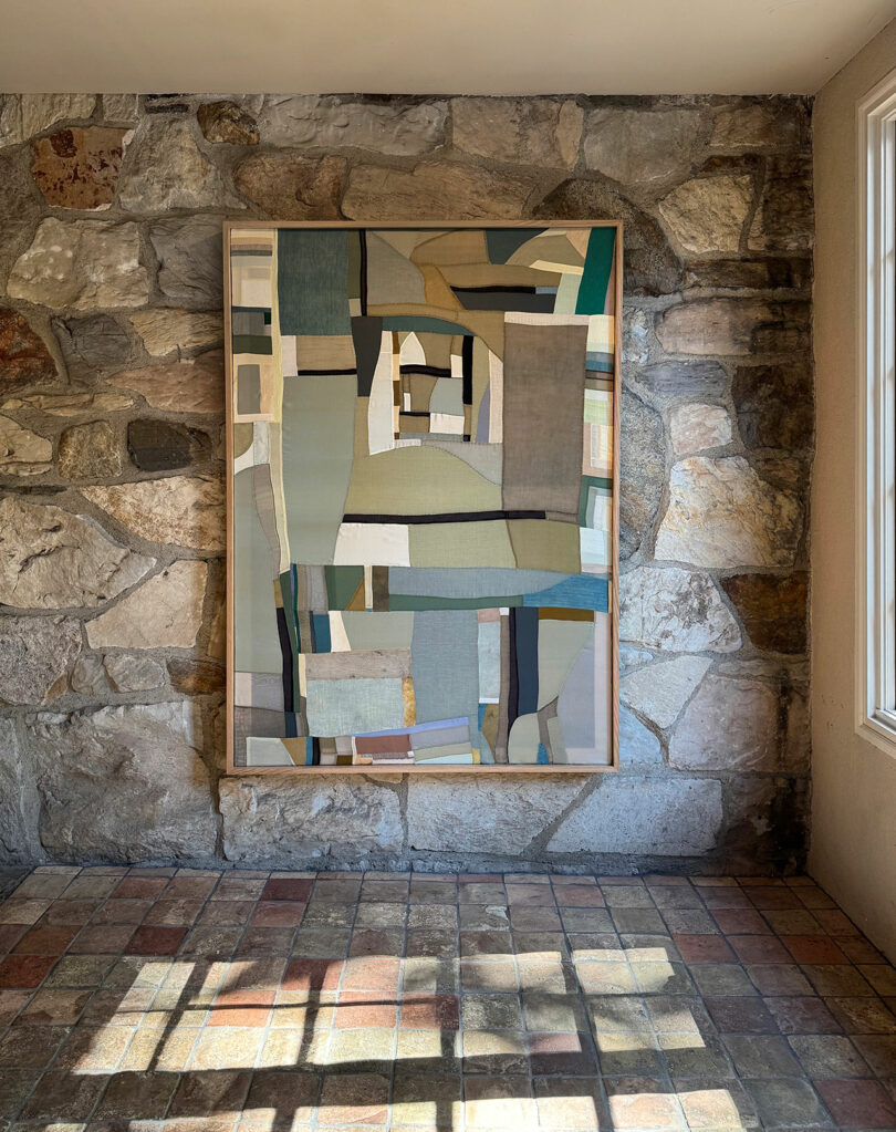 A geometric abstract painting by Kiva Motnyk with muted colors hangs on a stone wall, as sunlight casts grid-like shadows across the tiled floor.