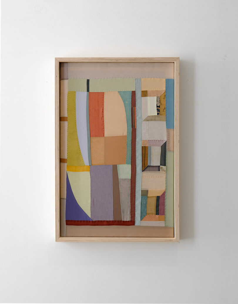 Framed abstract artwork by Kiva Motnyk featuring a patchwork of geometric shapes in muted colors—beige, blue, yellow, and orange—beautifully displayed on a plain white wall.