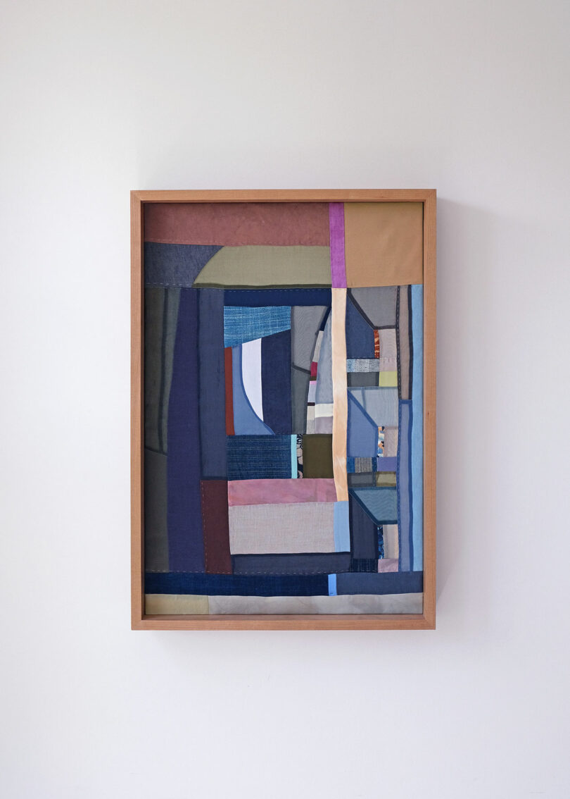Framed abstract textile artwork by Kiva Motnyk with geometric patchwork of various colored fabrics—including blue, brown, beige, purple, and gray—hanging on a plain white wall.