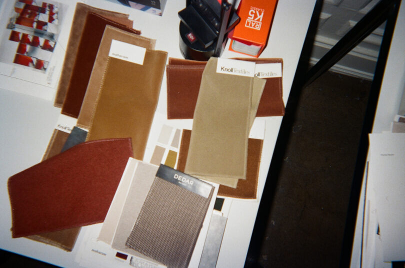 Assorted fabric swatches in shades of brown and red are spread out on a white table, alongside color sample cards and office supplies.