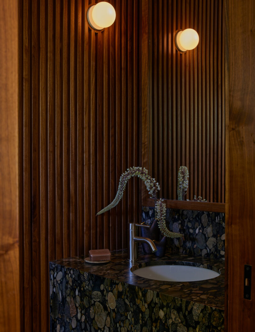 A bathroom with vertical wood panel walls, a stone-patterned counter with a sink, a modern faucet, a soap bar, and two round wall lights above the mirror.