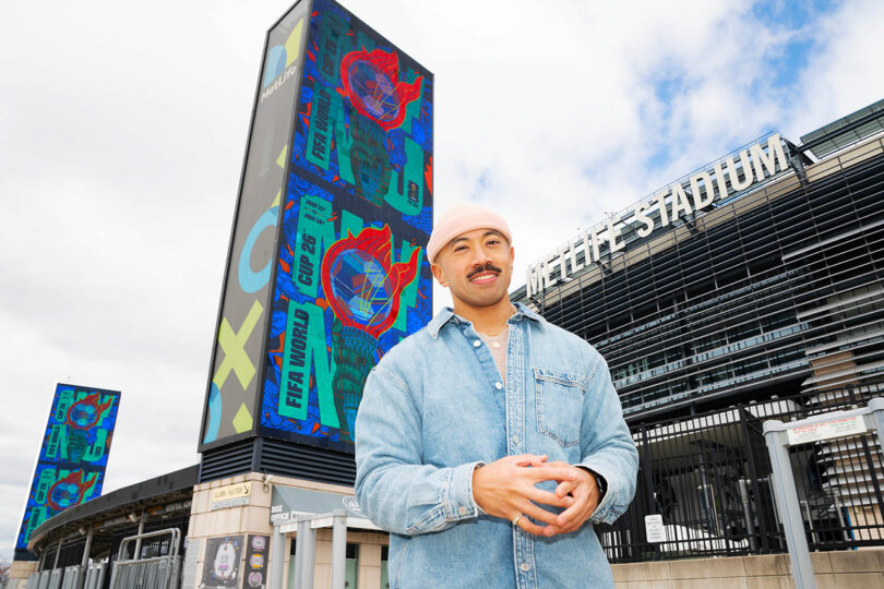 A person in a denim shirt stands in front of MetLife Stadium, with colorful digital FIFA World Cup 26 banners displayed outside.