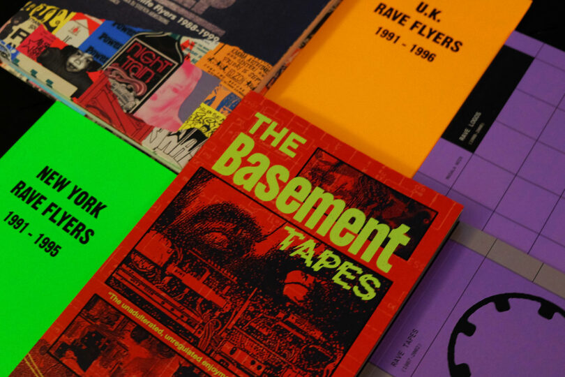 A closeup of colorful books and booklets, including “The Basement Tapes,” “NEW YORK RAVE FLYERS 1991-1995,” and “U.K. RAVE FLYERS 1991-1996,” arranged on a table.