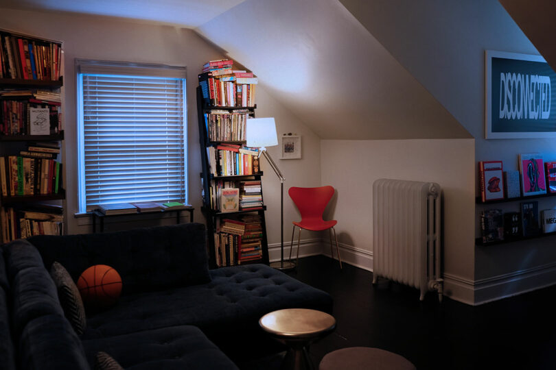 A cozy attic room with a sectional sofa, bookshelves filled with books, a red chair, a radiator, a window with blinds, and framed artwork on the wall.