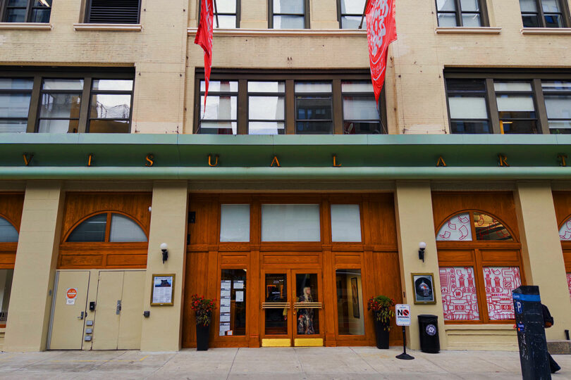 Street-level view of a building entrance with large wooden double doors, “VISUAL ARTS” signage above, and decorative red flags; window art and potted plants are visible.