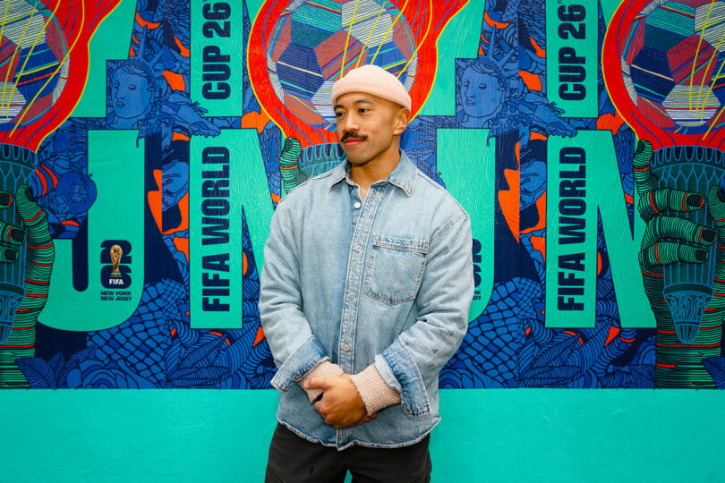 A person in a denim jacket and pink beanie stands in front of a colorful mural with "FIFA World Cup 26" text.