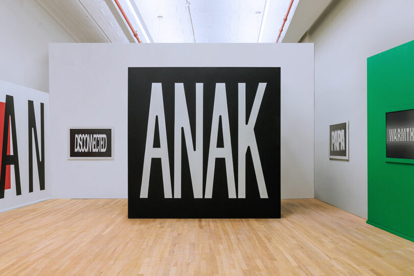 A gallery space with a large black box in the center featuring the word "ANAK" in bold white letters; framed text artworks are displayed on surrounding white and green walls.