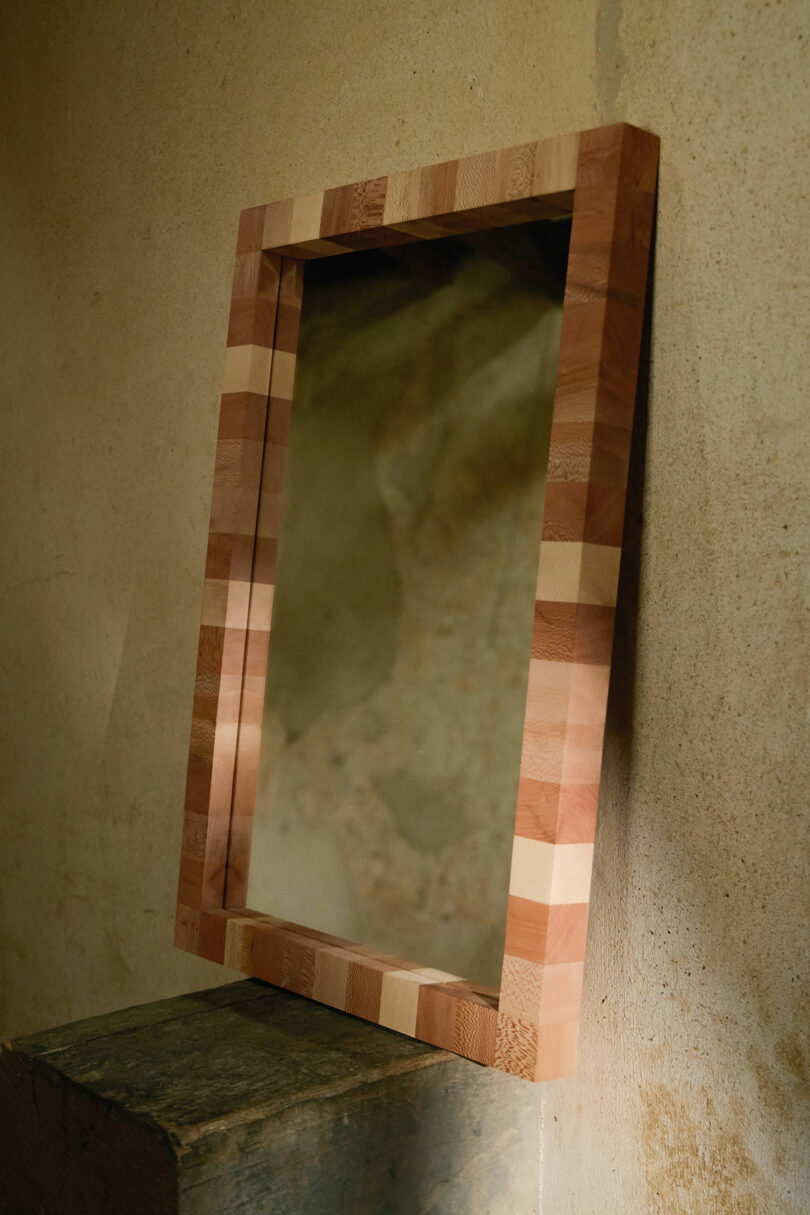 A rectangular mirror with a striped wooden frame is propped against a textured, beige wall and resting on a stone block.