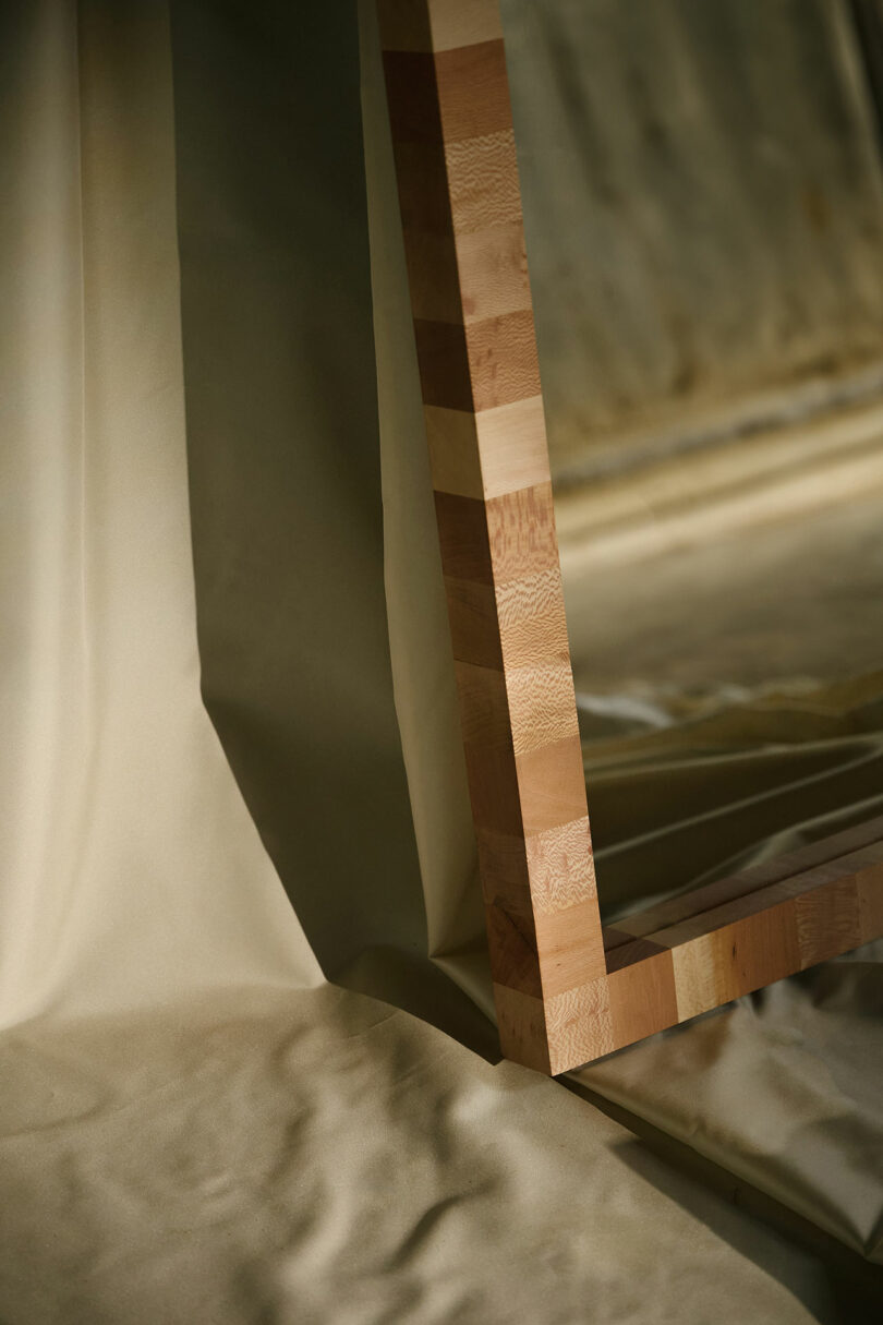 A close-up of a large wooden-framed mirror resting on beige fabric, reflecting soft natural light and muted surroundings.
