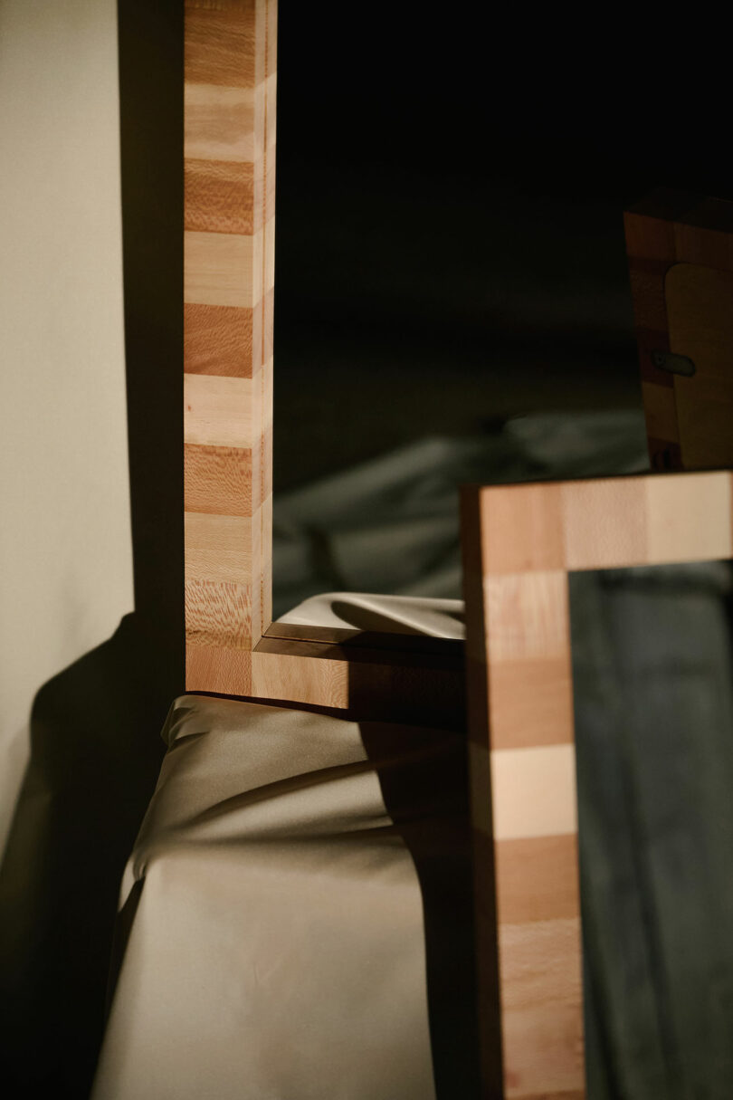 A close-up of a wooden frame with a striped pattern resting on a draped light-colored fabric, with shadows cast by the frame.
